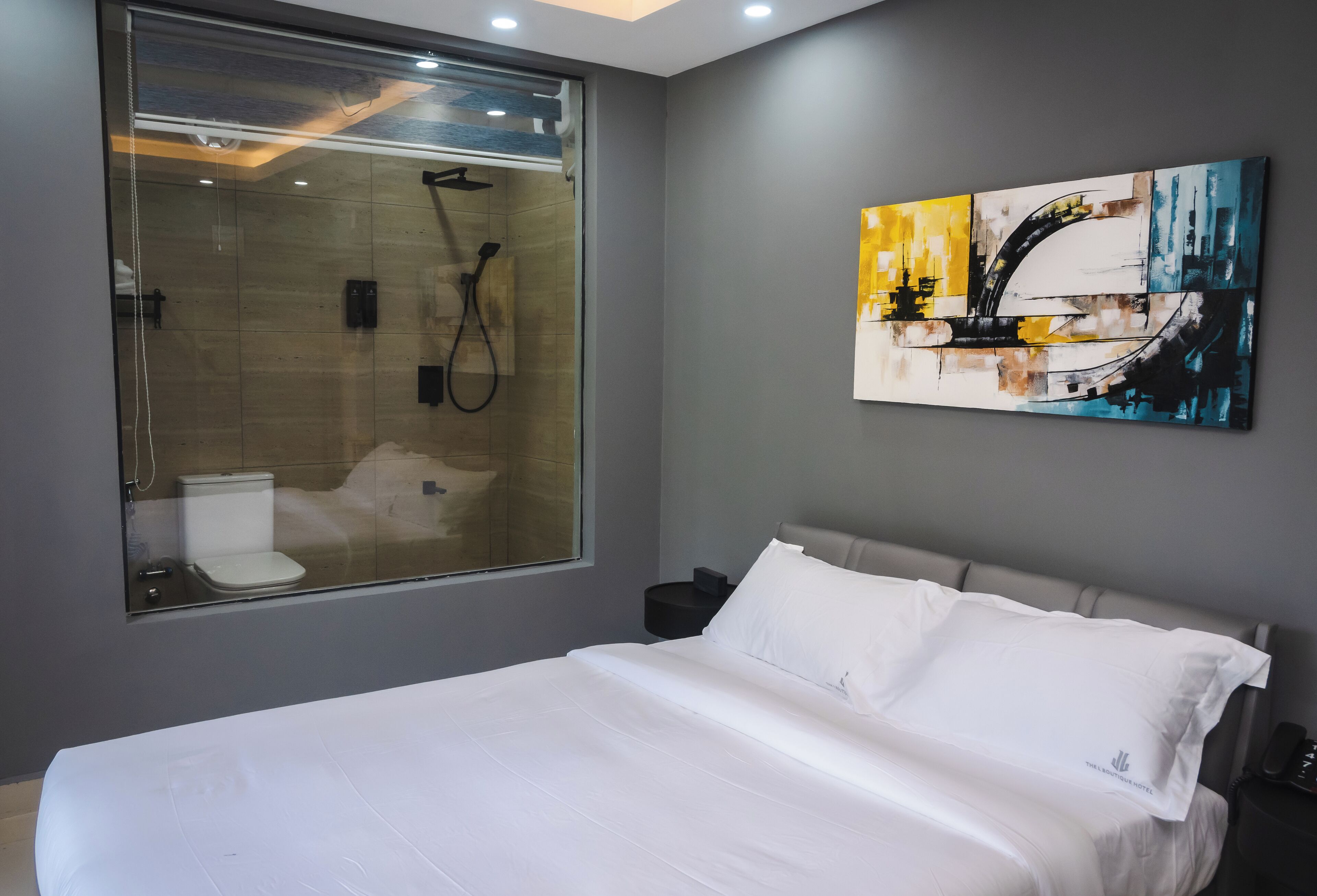 A modern hotel room with a large bed, a glass shower enclosure, a painting on the wall, and a small bedside table.