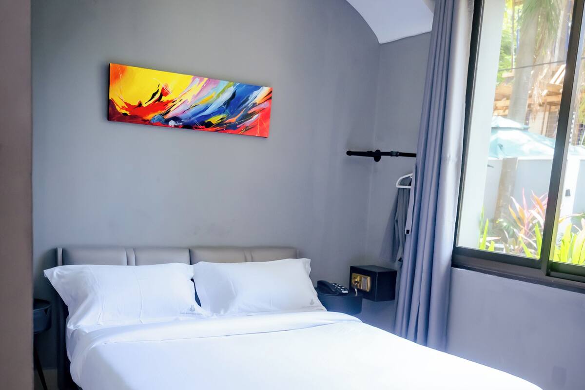 Ein ordentlich bezogenes Bett mit weißen Leinentüchern, ein farbenfrohes abstraktes Gemälde an der Wand und ein Paar schwarzer Hausschuhe auf dem Boden.