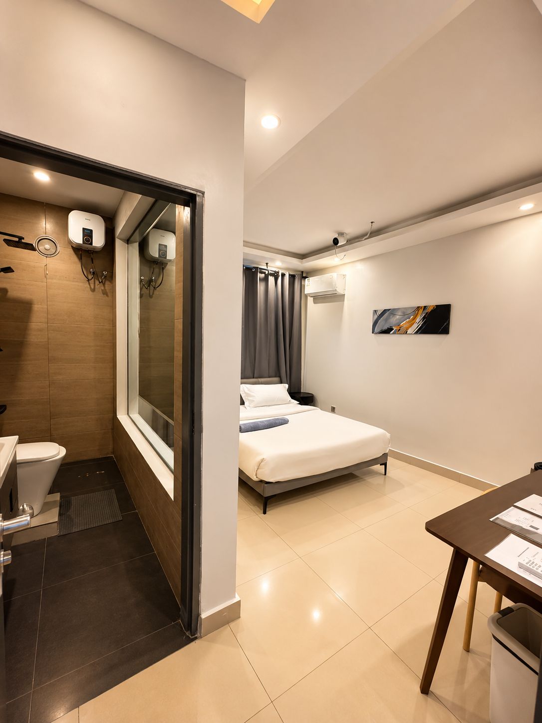 A modern hotel room with a shower, toilet, bed, and a small table with a trash bin.