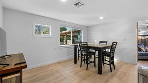 A dining area with a wooden table and chairs, a window with a view of greenery, and a white wall with a vent.