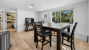 A modern dining area with a wooden table and chairs, a flat-screen TV, and a large window with a view of trees.