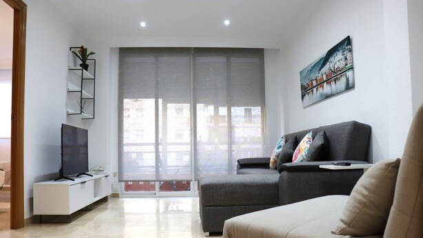 A modern living room with a grey sofa, a flat-screen TV, a white entertainment unit, and a large window with a view of buildings. - Apartment for groups with free parking  (Sevilla)