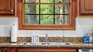 A kitchen with granite countertops, a stainless steel sink, wooden cabinets, and a window with a view of trees.