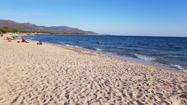 A sandy beach with clear blue water, a few people, and mountains in the background. - Spacious, quiet apartment close to the beach with magnificent views (Quartu Sant'Elena)