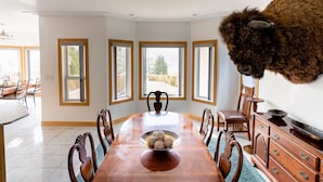 Une salle à manger avec une table en bois, des chaises et un saladier rempli de nourriture. Il y a des fenêtres avec des cadres en bois et une tête d’animal empaillée accrochée au mur.