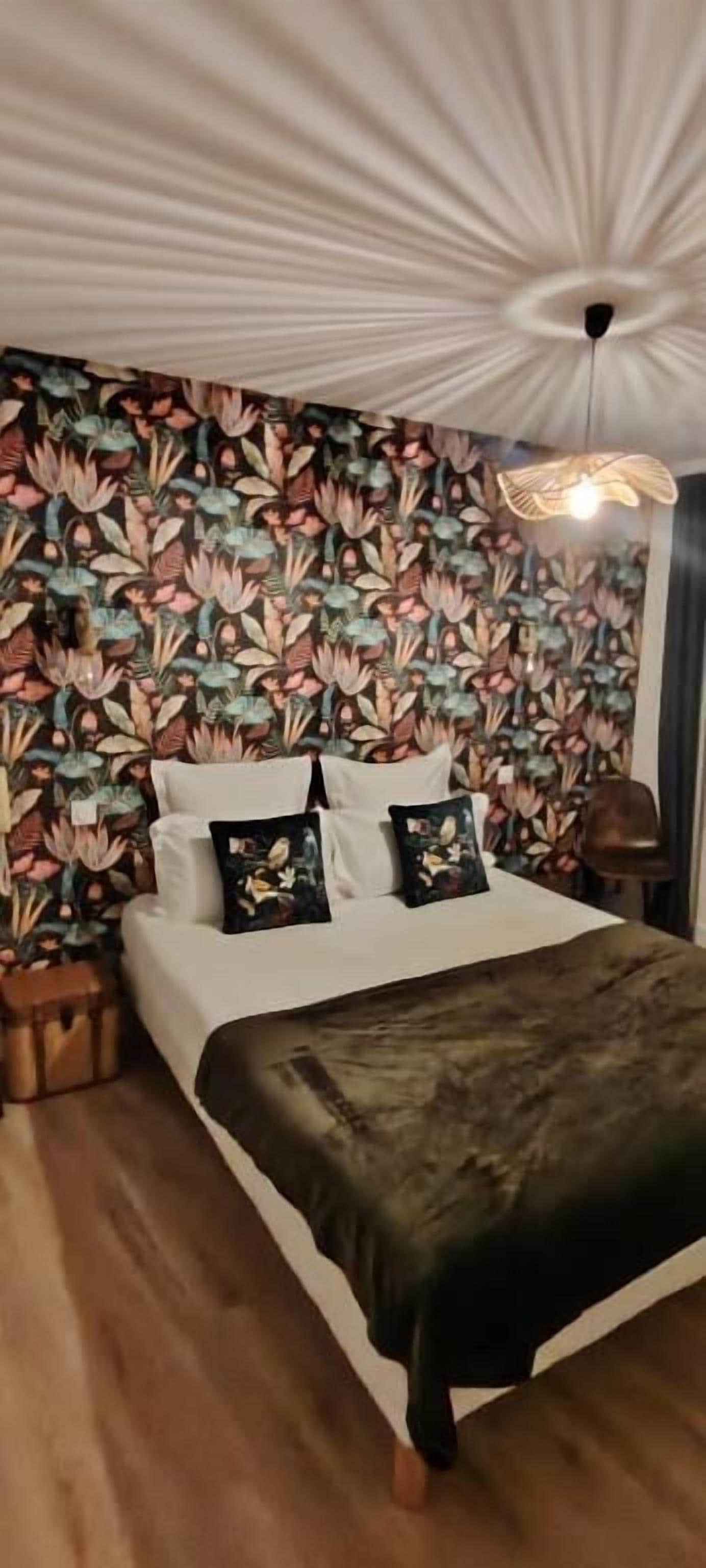 Un letto con lenzuola bianche e una coperta scura, una parete con motivo floreale e un lampadario.