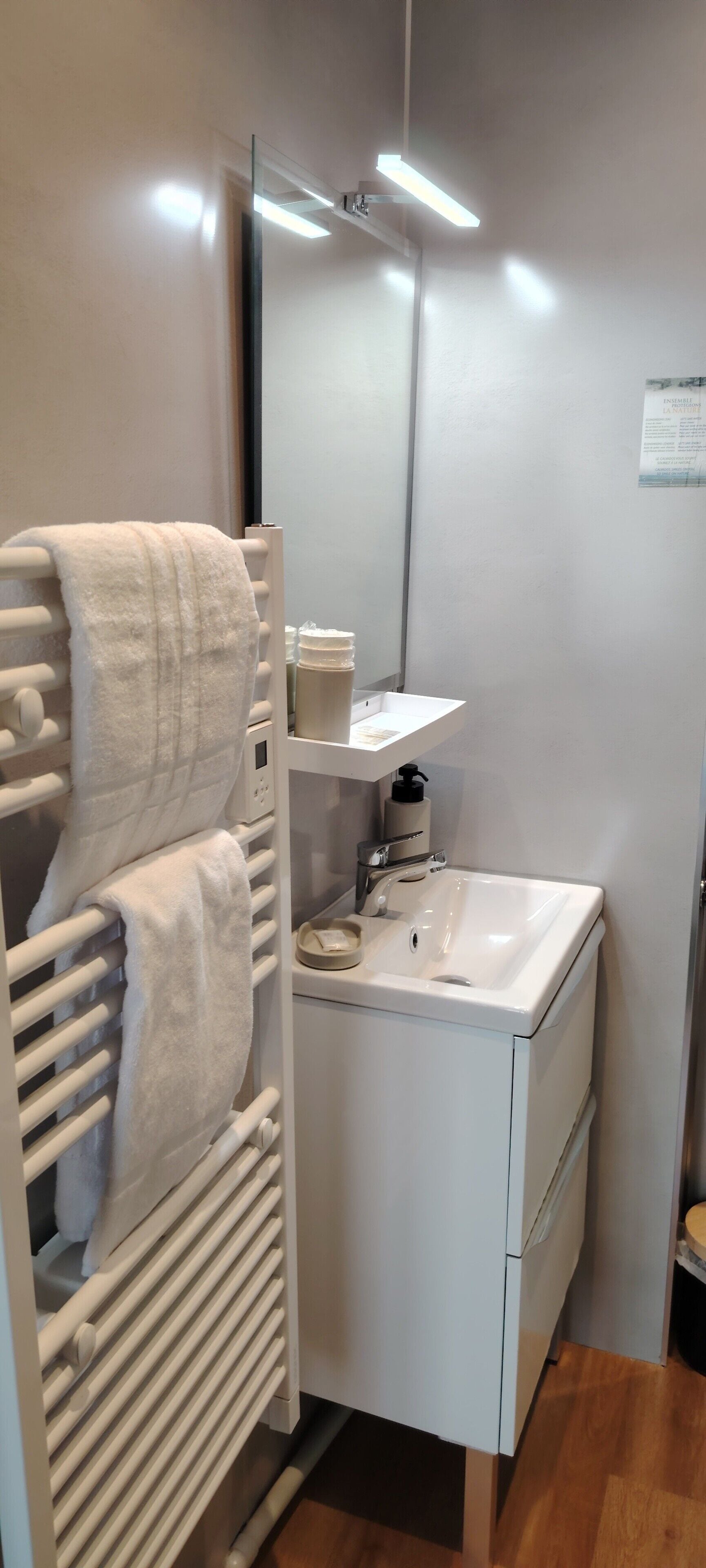 Un baño con un lavamanos blanco, un espejo, un perchero con toallas, una botella de jabón para manos y un estante pequeño con artículos de tocador.