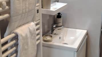 Un baño con un lavamanos blanco, un espejo, un perchero con toallas, una botella de jabón para manos y un estante pequeño con artículos de tocador.