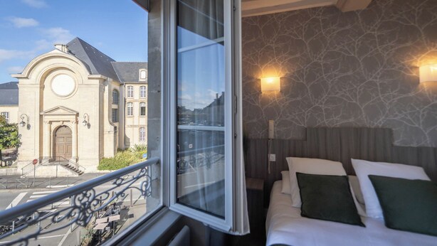 Una camera d'albergo con balcone, un letto con cuscini, un muro in pietra e vista su un edificio storico.