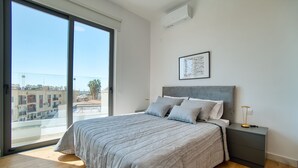 A bedroom with a large bed, a nightstand, a framed picture on the wall, and a view of buildings through a sliding door. - Arcadia 1 202 1 BED - One Bedroom Apartment, Sleeps 2 (Pafos)
