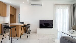 A modern living room with a flat-screen TV, a dining area with bar stools, and a kitchen with wooden cabinets. - Arcadia 1 202 1 BED - One Bedroom Apartment, Sleeps 2 (Pafos)