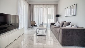 A modern living room with a flat-screen TV, a sofa, a coffee table, and a large window with sheer curtains. - Arcadia 1 202 1 BED - One Bedroom Apartment, Sleeps 2 (Pafos)