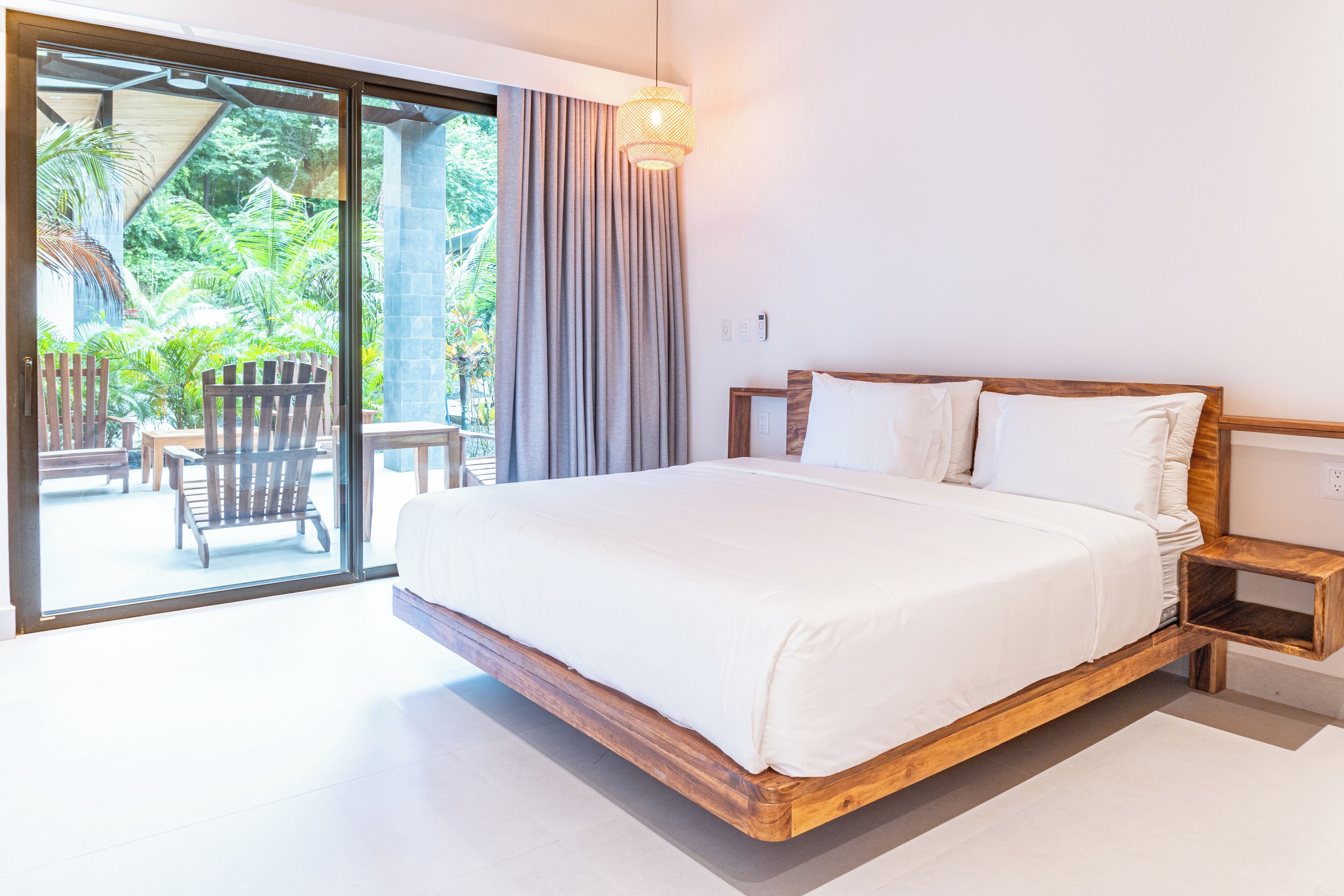 A bedroom with a large bed, wooden headboard, and a view of a patio with chairs and greenery.