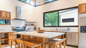Une cuisine moderne dotée d’un îlot central, de tabourets en bois, d’appareils électroménagers en acier inoxydable et d’une grande fenêtre offrant une vue sur la verdure.
