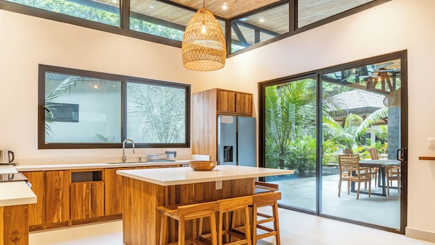 Une cuisine moderne dotée d’un grand îlot central, de tabourets de bar en bois et d’une grande fenêtre donnant sur un jardin tropical.