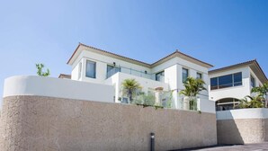 Une maison moderne à deux étages, avec une façade blanche, un mur de soutènement en pierre et un ciel d’un bleu éclatant.