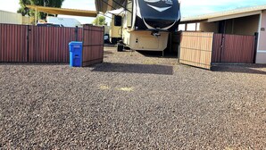 A recreational vehicle (RV) parked in a gravel lot with a covered area and a blue trash bin nearby. - Cozy RV with WiFi and HVAC in Glendale/North Phoenix Luxury Fifth wheel camper (Glendale)