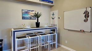 A reception area with a counter, three bar stools, a potted plant, a whiteboard, and a wall-mounted decorative item. - Harbor East Condominiums 414D by Ryson Vacation Rentals (Galveston)