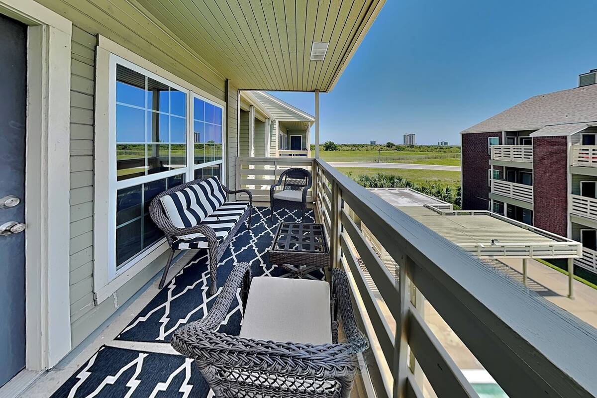 A balcony with wicker furniture, a patterned rug, and a view of a building and landscape.