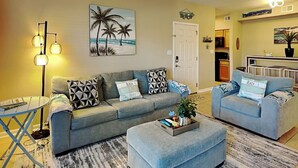 A living room with a grey sofa, a matching ottoman, a coffee table, and a floor lamp. There is a painting of palm trees on the wall and a door labeled "BEACH". - Harbor East Condominiums 414D by Ryson Vacation Rentals (Galveston)