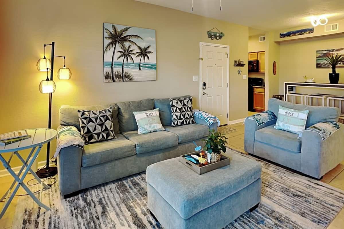A living room with a grey sofa, a matching ottoman, a coffee table, and a floor lamp. There is a painting of palm trees on the wall and a door labeled "BEACH".