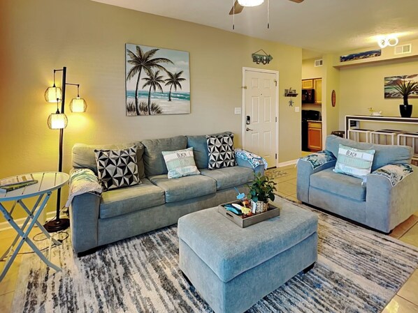 A living room with a grey sofa, a matching ottoman, a coffee table, and a floor lamp. There is a painting of palm trees on the wall and a door labeled "BEACH".