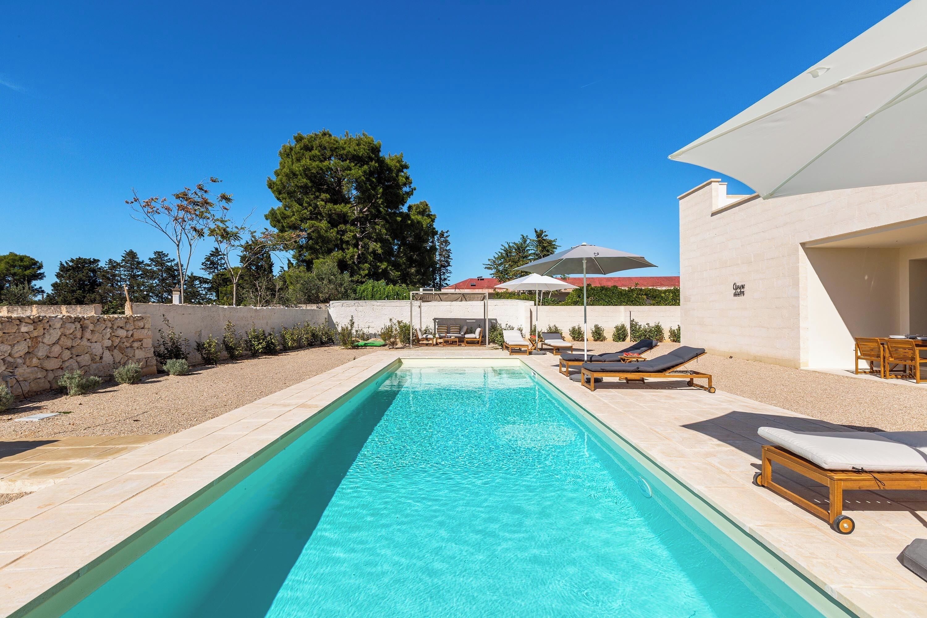 Piscina rectangular con sillones y sombrillas, rodeada por un muro de piedra y vegetación.