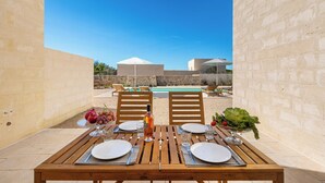 A wooden table set for dining with plates, cutlery, and a bottle of wine, overlooking a swimming pool and a stone wall. - Villa Li Santi - 2 by BarbarHouse (Santa Caterina)