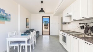 A modern kitchen with white cabinets, a marble countertop, and a built-in oven. There is a dining area with a white table and chairs, and a painting of a beach scene on the wall. - Villa Li Santi - 2 by BarbarHouse (Santa Caterina)