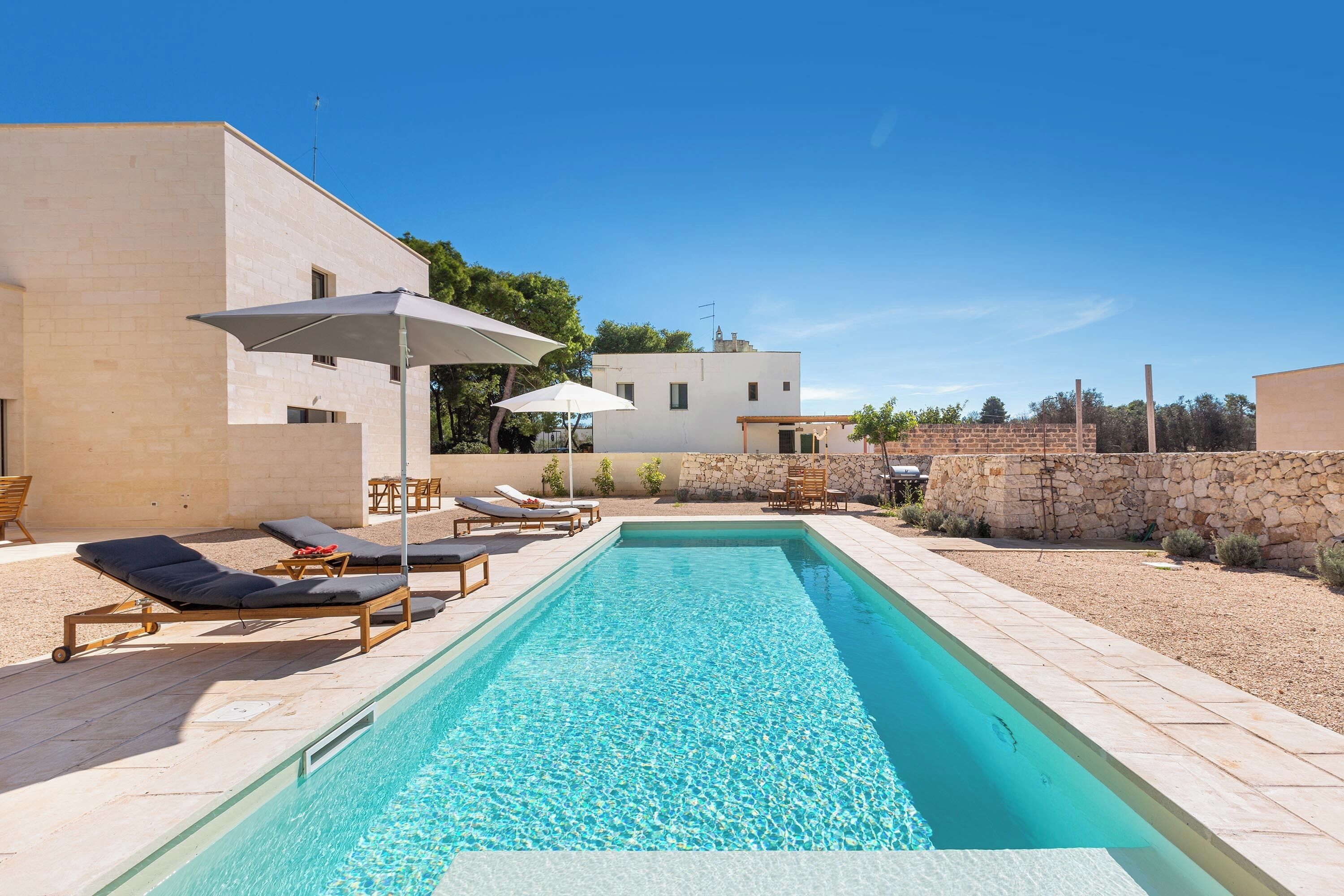 A rectangular swimming pool with lounge chairs and umbrellas, surrounded by a stone wall and a building with a flat roof.