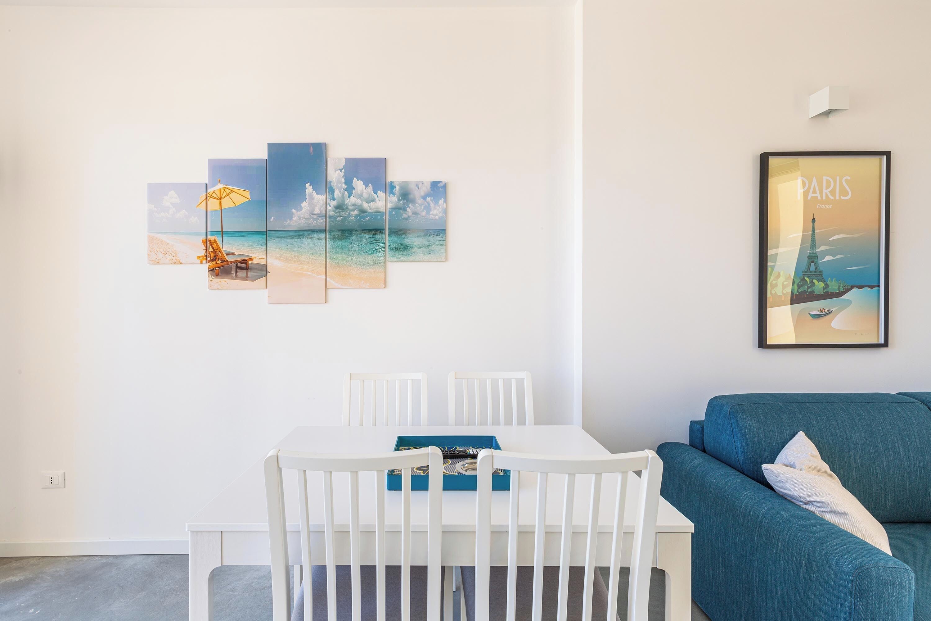 A room with a white dining table, white chairs, a blue sofa, and a wall with a beach scene and a Paris poster.