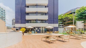 A modern building with a curved white wall, outdoor seating area with wicker chairs and umbrellas, and a playground.