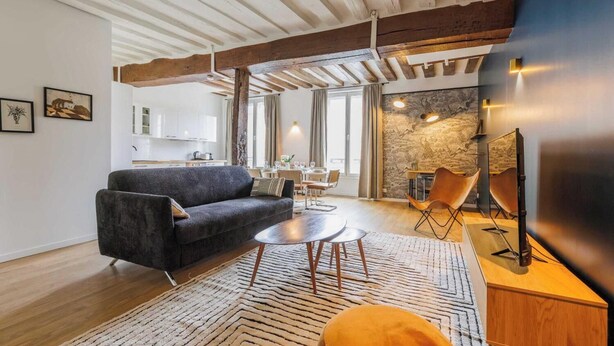 A modern living room with a sofa, coffee table, and TV stand. There is a kitchen in the background with white cabinets and a dining area with a table and chairs. - NESTLED IN THE LIVELY AND CHARMING NOTRE-DAME-DES-CHAMPS DISTRICT- RUE DE SEVRES (Paris)