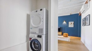 A compact, modern laundry room with a front-loading washer and dryer, a wooden floor, and a blue accent wall in the adjacent living area. - NESTLED IN THE LIVELY AND CHARMING NOTRE-DAME-DES-CHAMPS DISTRICT- RUE DE SEVRES (Paris)