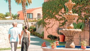 Un couple passe devant une fontaine en pierre à plusieurs niveaux, entourée de palmiers et d’un bâtiment à la façade en pierre.