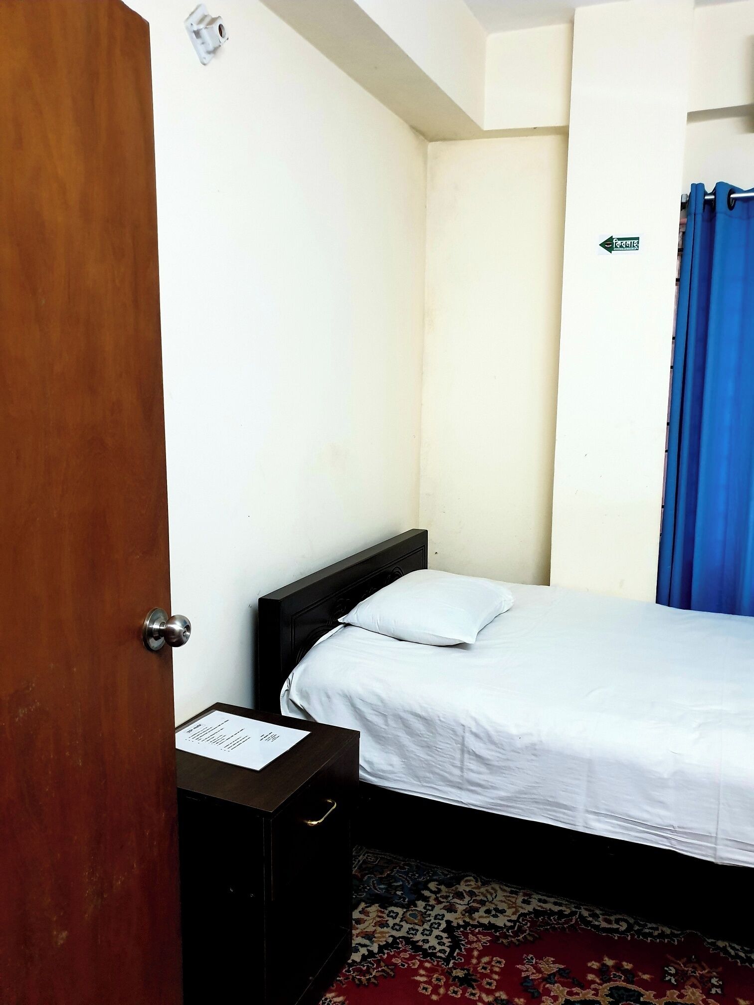 A bedroom with a wooden door, a bed with white bedding, a nightstand, a blue curtain, and a patterned rug.