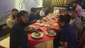 Un grupo de personas sentadas alrededor de una mesa con varios platos, en una habitación con un muro decorativo y una cortina estampada.