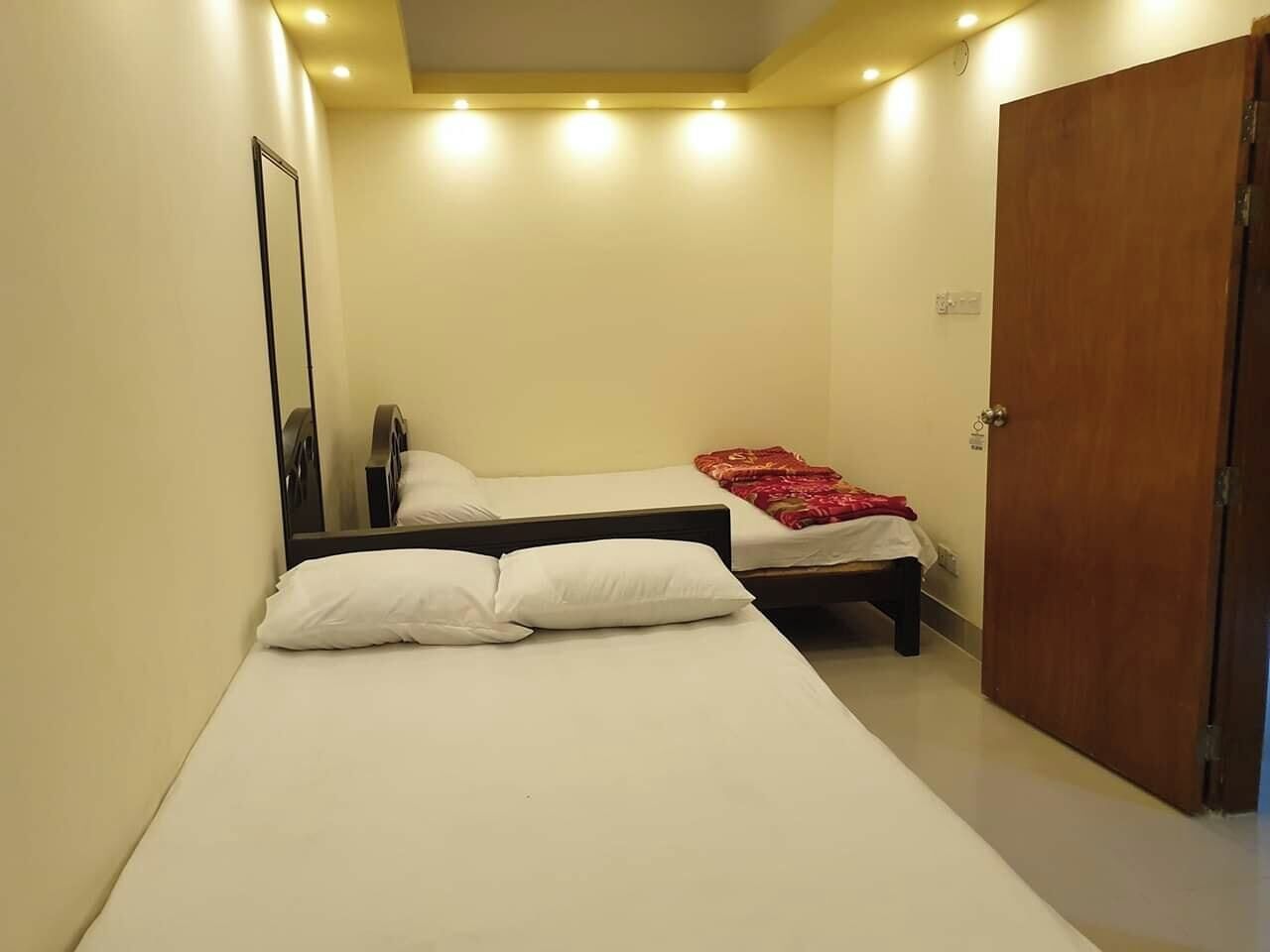 Une petite chambre bien éclairée, avec deux lits simples, une armoire en bois et un miroir au mur.