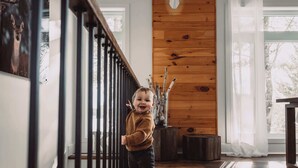 A child standing on a wooden staircase with a metal railing, a wooden wall with a deer head decoration, and a window with a view of trees.