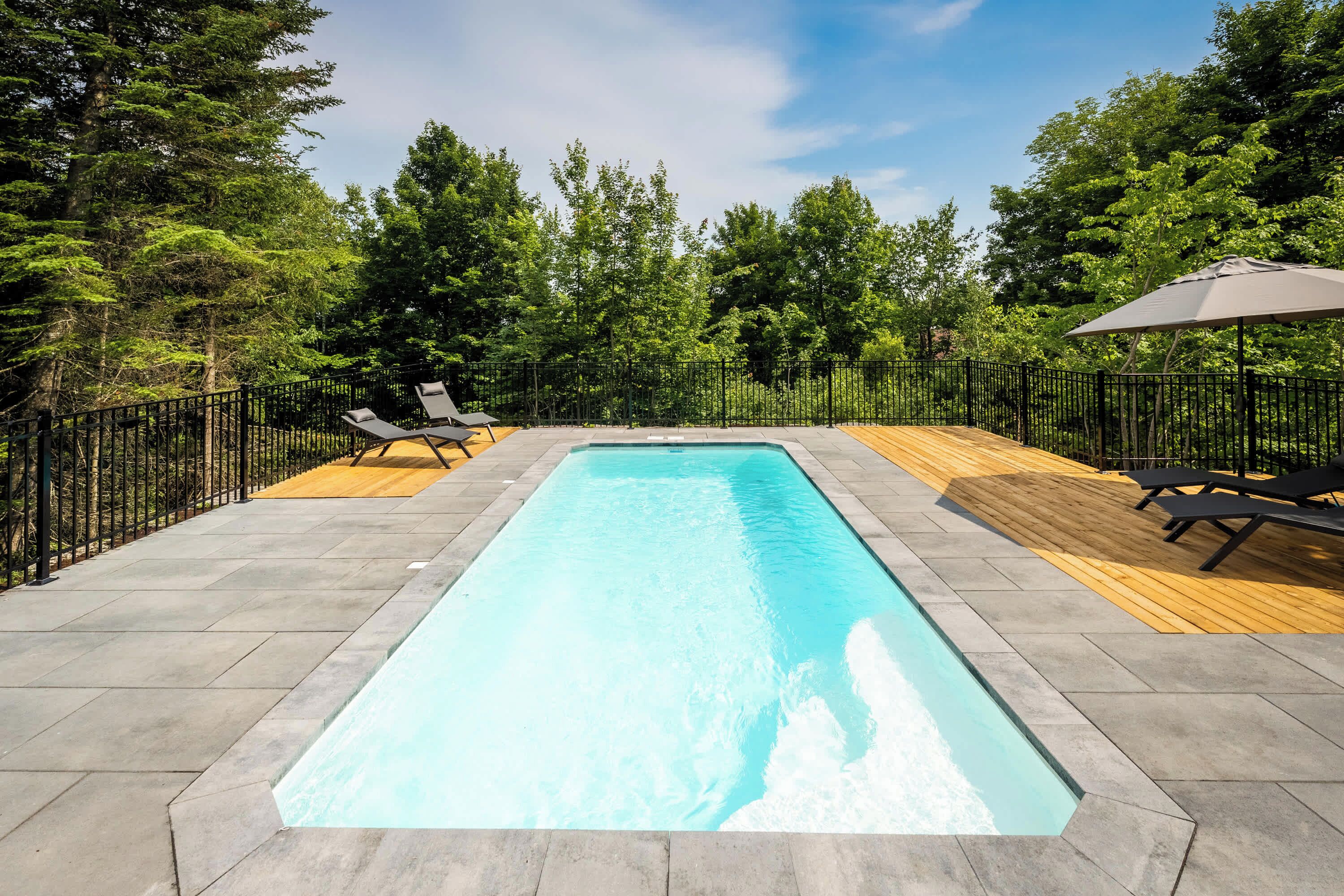 Ein rechteckiger Swimmingpool mit einer umlaufenden Holzterrasse, umgeben von einem schwarzen Metallzaun.