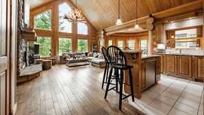 A spacious log cabin interior with a large living area, wooden floors, a stone fireplace, and a kitchen with wooden cabinets.