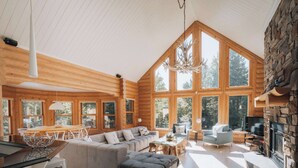 A spacious log cabin interior with a large living area, a fireplace, and multiple windows offering views of the surrounding forest.