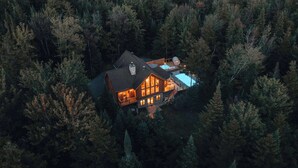 A large log cabin with a hot tub and a swimming pool, surrounded by dense forest.