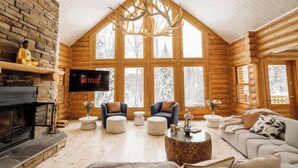 A log cabin interior with a stone fireplace, a large TV screen displaying 'NETFLIX', and a cozy seating area with sofas and ottomans.