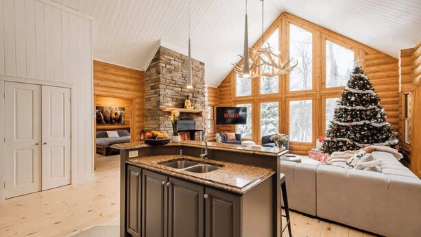 A log cabin interior with a kitchen island, stone fireplace, and a Christmas tree.