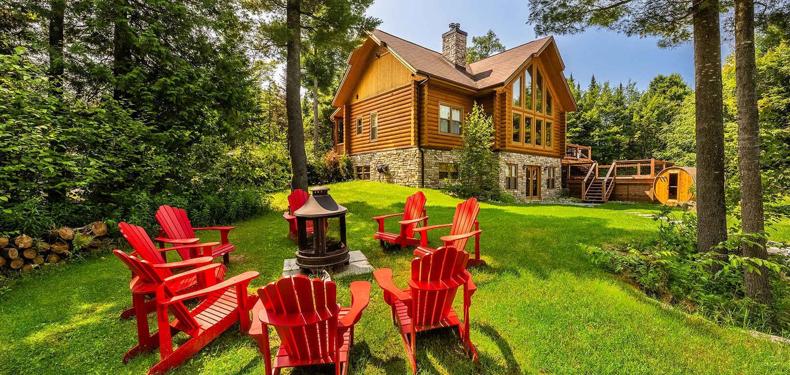 Una cabaña de troncos con cimientos de piedra, rodeada de un césped cuidado y sillones Adirondack rojos.