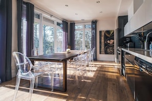 A modern kitchen with a wooden table and clear chairs, large windows with curtains, and a view of trees. - Chalet DesignPur on the Clermoustier Lake (Chertsey)