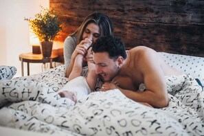 A couple lying on a bed with a patterned blanket in a room with a wooden wall and a small table with a lamp. - Chalet DesignPur on the Clermoustier Lake (Chertsey)