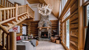 A log cabin interior with a stone fireplace, wooden staircase, and a large window. - Chalet Silver Fox at Fiddler Lake with Hot Tub, Sauna, and Private Pool (Mille-Isles)