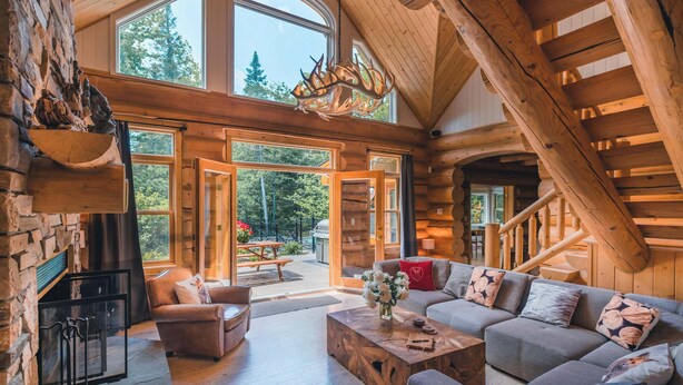 A spacious log cabin interior with a fireplace, large windows, a cozy seating area, and a wooden staircase. - Chalet Silver Fox at Fiddler Lake with Hot Tub, Sauna, and Private Pool (Mille-Isles)
